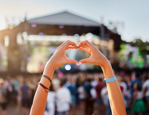Concertgoer forms heart shape with hands at beachside music festival, crowd enjoys live performance in sunset, love symbol gesture at summer event, youth celebration of art outdoor entertainment.
