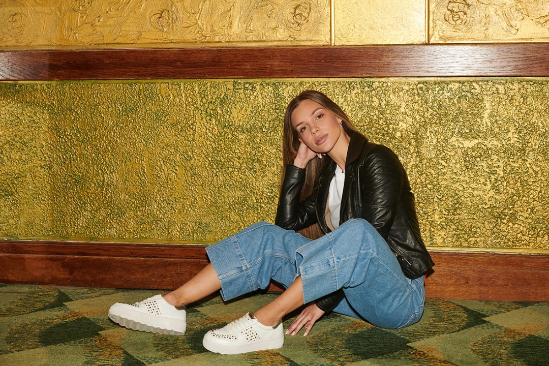 Woman poses wearing jeans, a leather jacket and metallic trainers